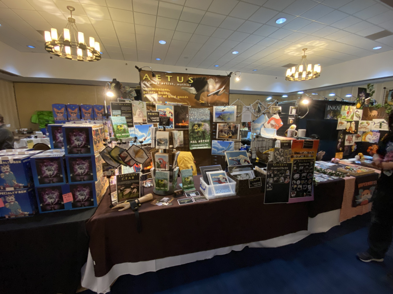 Convention display showing prints, masks, buttons, stickers, and books for sale.